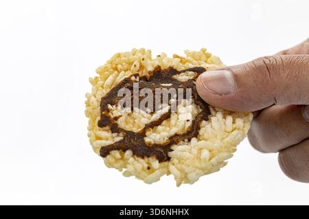 Gros plan d'une main tenant un gâteau de riz soufflé rond recouvert de sucre de canne mijoté foncé sur un fond blanc, mettant en valeur la texture du snack et le fl Banque D'Images