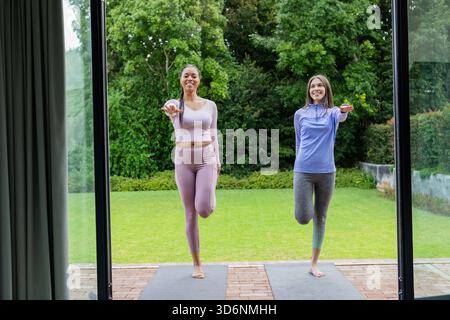 Amies féminines dans des vêtements d'entraînement souriant tout en équilibrant dans la pose d'arbre sur des tapis de yoga sur le patio Banque D'Images