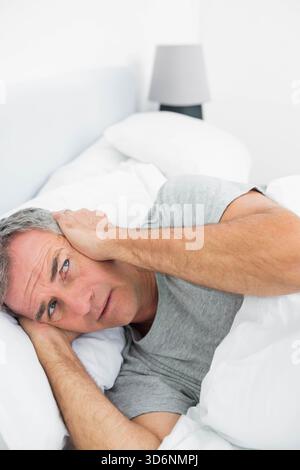 Homme mûr couché dans la chambre sur le lit avec oreiller pressant la main à l'oreille, portant un T-shirt gris. Minimaliste, monochrome, neutre, agité, anxieux, éveillé, Banque D'Images