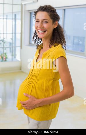 Femme enceinte debout dans le studio ensoleillé berçant bosse portant moutarde top pantalon léger anneau de mariage. Attendu, maternité, joyeux, lumineux, jaune, min Banque D'Images