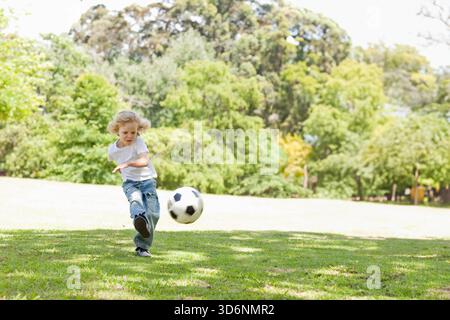 Garçon enfant frappant un ballon de football noir-blanc sur la pelouse gazonnée du parc portant un Jean T-shirt blanc et des chaussures. Jeune, à l'extérieur, espace vert, mouvement, sport, t Banque D'Images