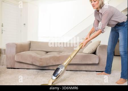 Femme d'âge moyen aspirant la moquette à la maison dans un chemisier beige et un Jean à l'aide d'un aspirateur doré, espace de copie. Nettoyant, sans fil, peluche, ensoleillé, neutre, minimal Banque D'Images