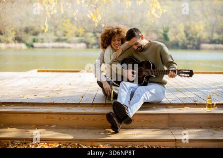 Un couple joyeux est assis sur une terrasse en bois au bord d'un lac serein, partageant des sourires tandis que l'un caresse une guitare et l'autre écoute, entouré de feuilles d'automne. Banque D'Images