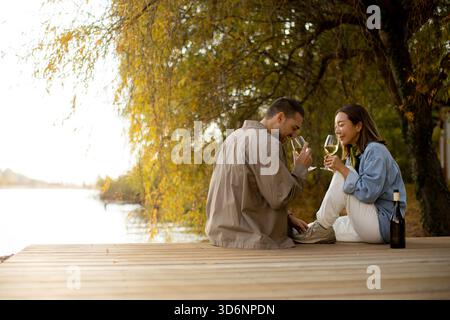 Un couple partage un moment joyeux au bord du lac tranquille au coucher du soleil, sirotant du vin et savourant la compagnie de l'autre sous les arbres. Banque D'Images
