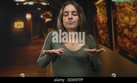 Femme avec les paumes vers le haut et les yeux fermés dans la galerie d'art du bâtiment, portant une chemise verte et un collier de coeur ; réflexion calme. Banque D'Images