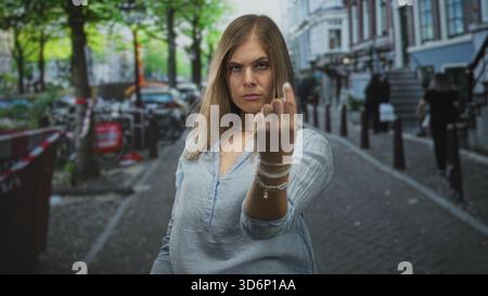 Jeune femme blonde se tient au milieu des vélos garés sur la rue pavée étroite retournant le majeur ; défi. Banque D'Images