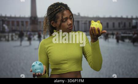 Femme afro-américaine tenant deux tirelires dans le bâtiment piazza, mains visibles en regardant une tirelire jaune ; sauver l'incertitude de choix. Banque D'Images