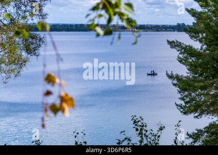Une scène sereine au bord du lac avec une eau bleue calme encadrée par des branches d'arbres au premier plan. Au loin, un petit bateau avec deux personnes flotte tranquillement Banque D'Images