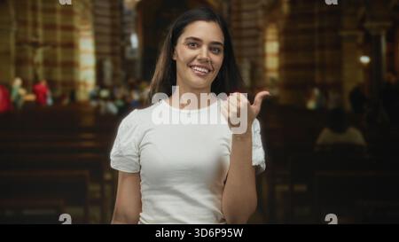 Femme hispanique jeune pointe pouce à côté à l'intérieur du bâtiment de l'église, souriant et debout par des bancs en bois sous la lumière intérieure chaude ; joie accueillante. Banque D'Images