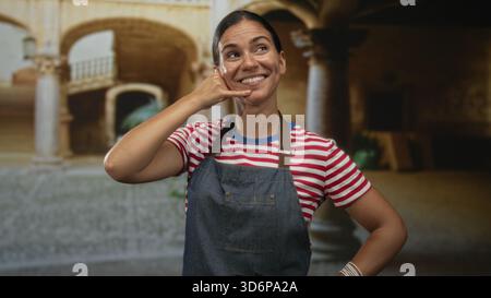 Femme fait appel à moi geste de la main tout en souriant dans la vieille cour du bâtiment portant tablier et chemise rayée ; salutation ludique. Banque D'Images