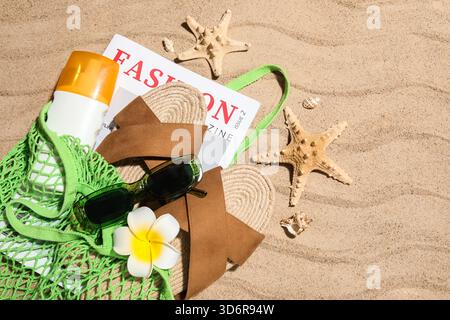 Sac à ficelle avec bouteille de crème solaire, magazine et accessoires de plage sur sable comme fond, gros plan Banque D'Images
