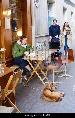 Paris, France - mars 2025 : une femme est assise à une petite table pour manger, tandis qu'un couple avec une poussette se tient à proximité. Un chien se détend sur le sol à l'extérieur d'un Banque D'Images