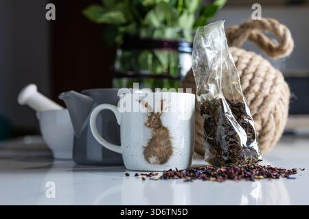 Flash allumé gros plan du thé aux fruits en vrac renversé d'un paquet ouvert sur le comptoir de cuisine en marbre avec tasses en céramique, théière, pilon et mortier la nuit Banque D'Images