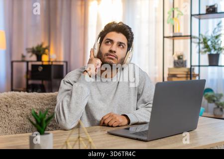 Jeune homme indien portant des écouteurs sans fil travaillant sur un ordinateur portable, semblant concentré tout en écoutant un livre audio. Le gars hispanique est assis sur le canapé à table dans le salon à la maison, profondément engagé dans sa tâche. Banque D'Images