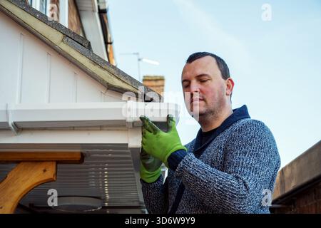 Homme sur une échelle nettoyant les feuilles d'automne tombées et la mousse de la gouttière du toit de la maison. Entretien de la maison, nettoyage des gouttières Banque D'Images