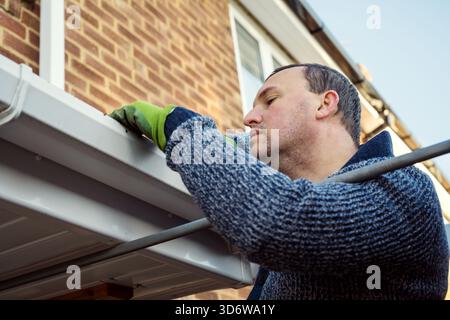 Homme sur une échelle nettoyant les feuilles d'automne tombées et la mousse de la gouttière du toit de la maison. Entretien de la maison, nettoyage des gouttières Banque D'Images