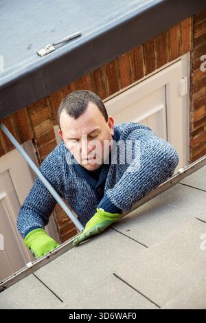 Homme sur une échelle nettoyant les feuilles d'automne tombées et la mousse de la gouttière du toit de la maison. Entretien de la maison, nettoyage des gouttières Banque D'Images
