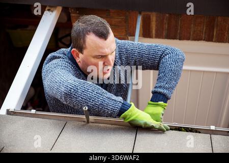 Homme sur une échelle nettoyant les feuilles d'automne tombées et la mousse de la gouttière du toit de la maison. Entretien de la maison, nettoyage des gouttières Banque D'Images