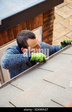 Homme sur une échelle nettoyant les feuilles d'automne tombées et la mousse de la gouttière du toit de la maison. Entretien de la maison, nettoyage des gouttières Banque D'Images