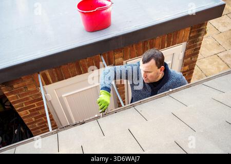 Homme sur une échelle nettoyant les feuilles d'automne tombées et la mousse de la gouttière du toit de la maison. Entretien de la maison, nettoyage des gouttières Banque D'Images
