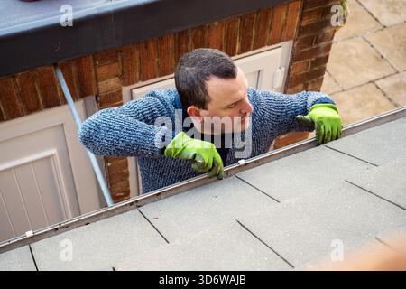 Homme sur une échelle nettoyant les feuilles d'automne tombées et la mousse de la gouttière du toit de la maison. Entretien de la maison, nettoyage des gouttières Banque D'Images