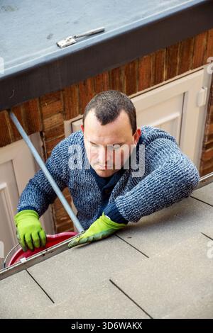 Homme sur une échelle nettoyant les feuilles d'automne tombées et la mousse de la gouttière du toit de la maison. Entretien de la maison, nettoyage des gouttières Banque D'Images