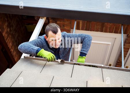 Homme sur une échelle nettoyant les feuilles d'automne tombées et la mousse de la gouttière du toit de la maison. Entretien de la maison, nettoyage des gouttières Banque D'Images