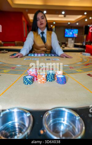 Piles de jetons de casino colorés sur la table de baccarat avec croupier flou en arrière-plan montrant l'action de jeu, le risque de pari, la chance et l'atmospher de jeu Banque D'Images