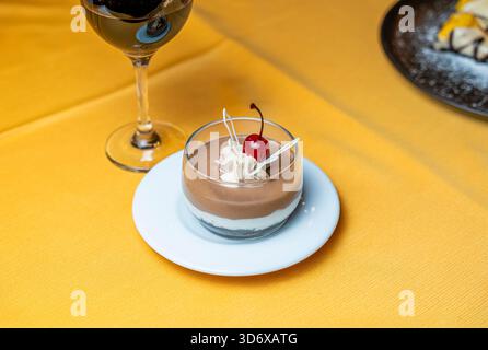 Dessert panna cotta au chocolat garni de cerise, crème fouettée et décoration au chocolat blanc servi dans une tasse en verre sur une assiette blanche sur un caillot jaune Banque D'Images