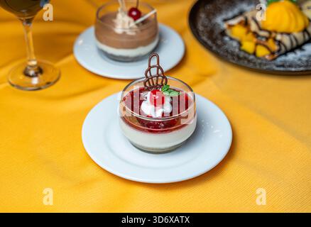 Dessert à la panna cotta aux baies garni de cerise, crème fouettée, menthe et décoration chocolat servi dans une tasse en verre sur une assiette blanche sur un tissu jaune. Banque D'Images