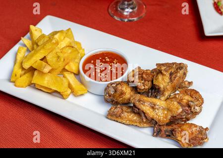 Ailes de poulet frites croustillantes avec frites dorées et sauce piquante sur une assiette blanche pour les promotions culinaires. Banque D'Images