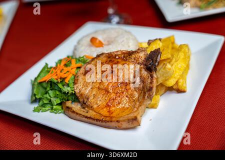 Côtelette de porc grillée servie avec riz blanc, salade fraîche et yucca frit sur une assiette blanche sur une nappe rouge pour le menu ou la promotion des aliments. Banque D'Images
