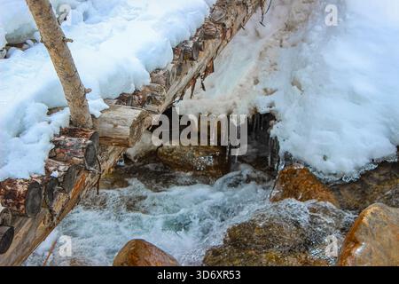 Une photographie d'un vieux pont en bois couvert de neige au-dessus d'un ruisseau de montagne qui coule rapidement. Les glaçons pendent des bords, créant une atmosphère hivernale Banque D'Images