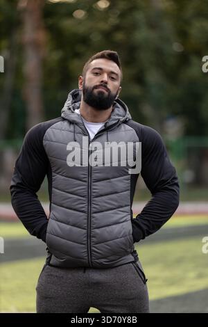athlétique homme se tient debout sur un terrain de sport portant une veste ajustée et regardant avec confiance l'appareil photo. Le cadre présente un champ vert vibrant et des arbres en arrière-plan. Banque D'Images