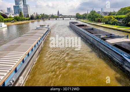 Deux grandes péniches de fret passent le long de la rivière main avec l'horizon de la ville et la flèche lointaine de l'église visible sous un ciel lumineux. Banque D'Images