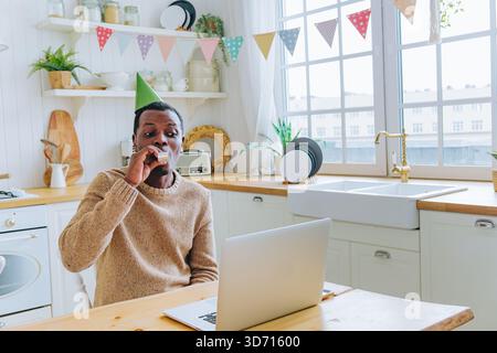 Homme afro-américain dans la cuisine portant un chapeau d'anniversaire, soufflant joyeusement une corne de fête lors d'un appel vidéo d'anniversaire virtuel sur son ordinateur portable, se connectant avec ses proches à distance Banque D'Images