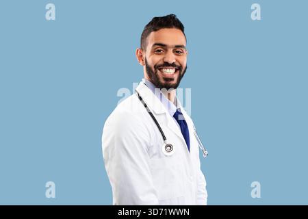 Jeune médecin masculin souriant avec confiance sur un fond bleu, mettant en valeur le professionnalisme et l'accessibilité Banque D'Images