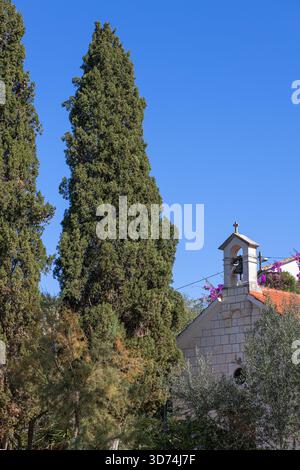 Une scène côtière tranquille comprend une chapelle en pierre pittoresque encadrée par de grands cyprès et une végétation luxuriante, capturant le charme rustique et la nostalgie. Banque D'Images