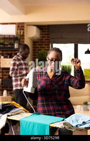 Femme afro-américaine dansant et repassant les vêtements dans le salon, profitant des tâches ménagères du week-end. Black Housewoman tient le fer électrique pour presser les chemises froissées, rendant le ménage amusant et animé à la maison. Banque D'Images