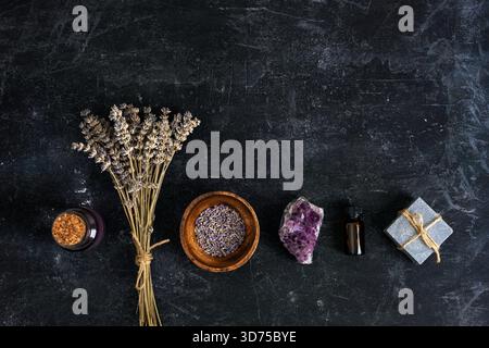 Fleurs de lavande séchées, bol en bois rempli de lavande, cristal d'améthyste, bouteille avec huile essentielle et savon fait main sur fond noir texturé. Banque D'Images