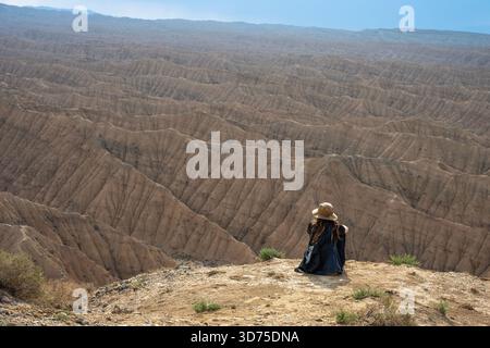 Voyageur seul portant un chapeau et un sac à dos est assis sur le bord d'une haute falaise, regardant au-dessus du paysage texturé expansif et profondément érodé de l'Ak- Banque D'Images