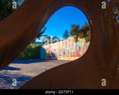 Murale colorée « Saludos from Cloverdale » encadrée à travers une sculpture en métal rouillé sous un ciel bleu vif. Banque D'Images