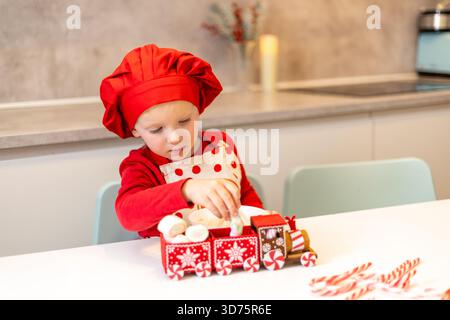 Un enfant joyeux dans un chapeau de chef rouge et un tablier place soigneusement des bonbons sur un train jouet. La cuisine est décorée pour les vacances, créant un atmos chaleureux Banque D'Images