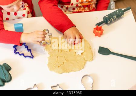 Les enfants déploient de la pâte à biscuits sur une table, en utilisant des emporte-pièces colorés pour créer des formes festives. L'atmosphère est joyeuse, avec un accent sur ho Banque D'Images