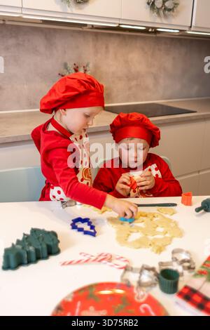 Deux enfants cuisent joyeusement des biscuits de Noël dans une cuisine chaleureuse et décorée. Ils portent des chapeaux de chef rouges assortis et semblent concentrés car ils utilisent co Banque D'Images