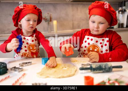 Deux enfants portent une tenue festive tout en cuisinant joyeusement des biscuits dans une cuisine chaude. Ils utilisent des emporte-pièces et de la pâte sur la table, mettant en valeur un joyeux A. Banque D'Images