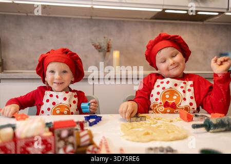 Deux enfants portant des chapeaux et des tabliers rouges vifs préparent joyeusement des biscuits. Ils sont entourés de pâte à biscuits et de décorations festives dans un kitch moderne Banque D'Images