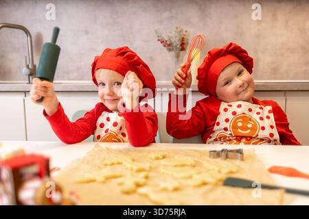 Deux enfants sourient joyeusement dans une cuisine lumineuse tout en cuisinant des biscuits. Ils portent des chapeaux rouges joyeux et des tabliers, montrant leur excitation pour le holid Banque D'Images