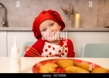 Un enfant joyeux vêtu d'un chapeau de chef rouge et d'un tablier crée des biscuits festifs dans une cuisine confortable. L'enfant tient un fouet et aime le processus de cuisson Banque D'Images