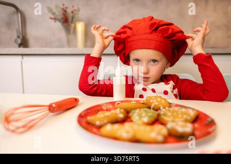 Un enfant joyeux dans un chapeau de chef rouge est assis à la table, prêt à décorer des biscuits colorés de vacances. La cuisine est lumineuse, créant une atmosphère joyeuse Banque D'Images